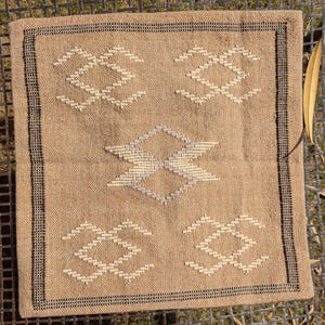 May include: Square beige pillow with geometric white and gray embroidered designs. The pillow has a black border and is placed on a metal surface. A dried leaf is on the right side of the pillow.