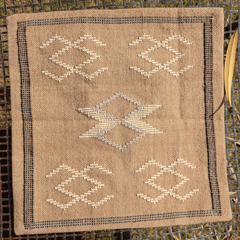 May include: Square beige pillow with geometric white and gray embroidered designs. The pillow has a black border and is placed on a metal surface. A dried leaf is on the right side of the pillow.