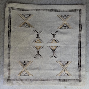 May include: Square off-white cushion with a geometric pattern. The cushion features a repeating design of hourglass and diamond shapes in black and gold. A black border frames the edges.