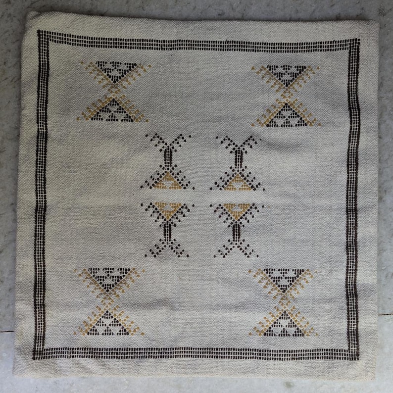 May include: Square off-white cushion with a geometric pattern. The cushion features a repeating design of hourglass and diamond shapes in black and gold. A black border frames the edges.