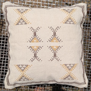May include: Square cream-colored pillow with a geometric pattern in brown and gold. The pillow has a textured surface and a brown border. The pattern consists of repeating shapes, adding a decorative touch to the pillow.