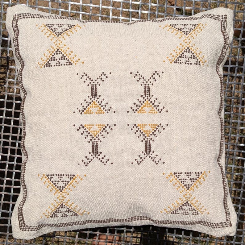 May include: Square cream-colored pillow with a geometric pattern in brown and gold. The pillow has a textured surface and a brown border. The pattern consists of repeating shapes, adding a decorative touch to the pillow.
