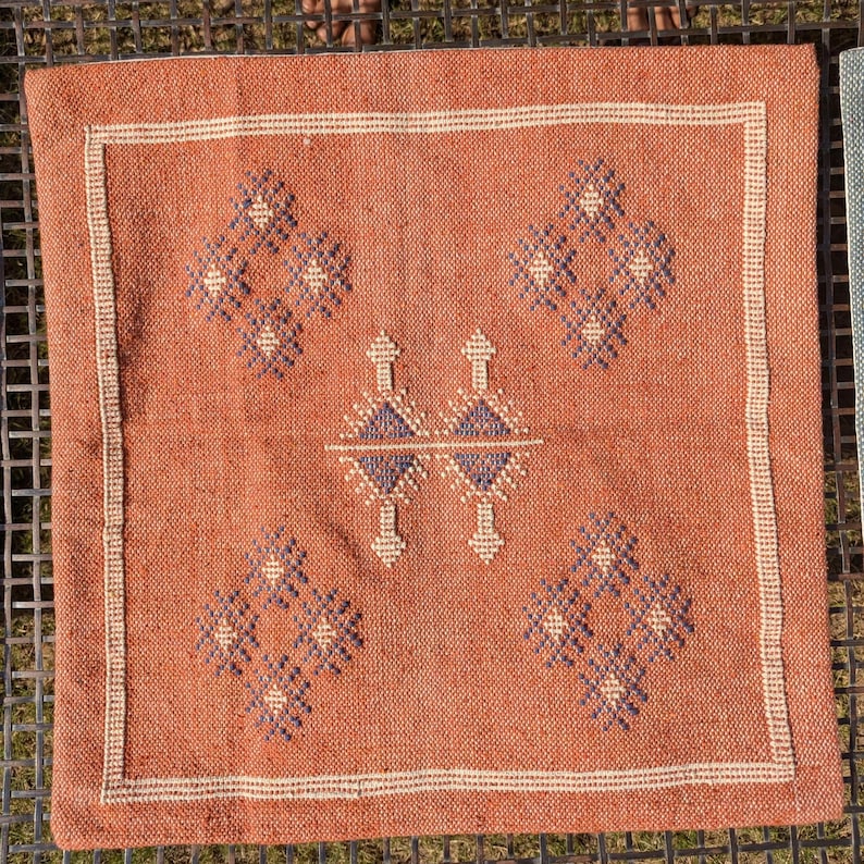 May include: Square terracotta-colored pillow with a geometric design. The pillow features a central diamond pattern flanked by four diamond-shaped motifs. The edges are bordered with a cream-colored trim.