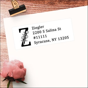 Könnte beinhalten: Ein weißes Adressetikett mit dem Namen "Ziegler" und Adressinformationen. Der Buchstabe "Z" ist mit einem floralen Design verziert. Eine schwarze und goldene Büroklammer befindet sich oben links. Eine rosa Blume befindet sich in der unteren linken Ecke.