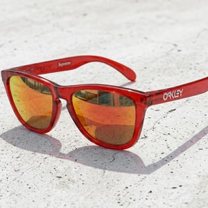 May include: Translucent red sunglasses with reflective orange-tinted lenses. The frame features the text Frogskins and Supreme printed on the inner temple, while the outer temple displays the white Oakley logo on the side of the arm.