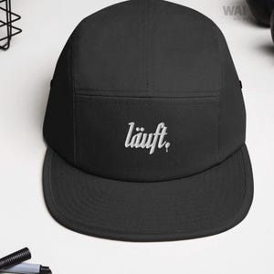 Może przedstawiać: Czarna czapka z pięcioma panelami i płaskim daszkiem. Czapka ma biały haft z napisem "läuft" z przodu. Czapka jest wykonana z wytrzymałego materiału i ma strukturalny design.