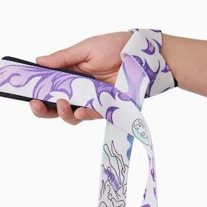 Puede incluir: Correas blancas para levantamiento de pesas con un diseño de llamas moradas y azules. Las correas se sostienen en una mano y se envuelven alrededor de la muñeca. El diseño incluye contornos negros y un pequeño logotipo circular.