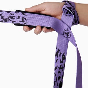 Purple anime lifting straps with black flame design being held. Heavy-duty powerlifting gear with character badge detail and secure loop construction for deadlifts and strength training.