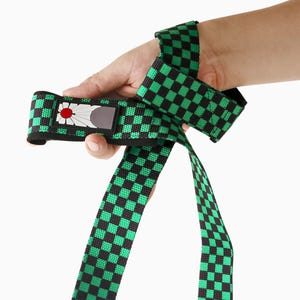 May include: Green and black chequered weightlifting wrist strap with a red and white emblem. The strap is held in a hand against a white background. The strap is designed to wrap around the wrist for support during weightlifting exercises.