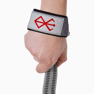 Op de afbeelding: Een polsband met een zilverkleurige patch met een rood symbool. De band heeft een zwarte en grijze rand en wordt om de pols vastgemaakt. Een grijze en zwarte band wordt in de hand gehouden.