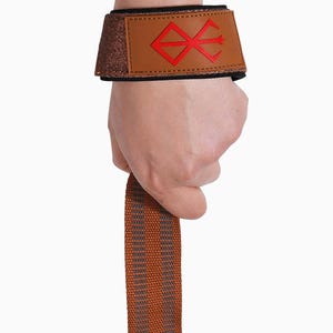 Op de afbeelding: Een bruine en zwarte polsband met een rood grafisch ontwerp. De polsband heeft een bruin leren patch met een rood symbool en een glinsterend bruin gedeelte. Een hand houdt de band vast, met een bruin en grijs gestreepte band.