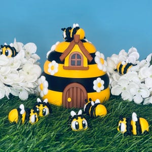 May include: A yellow and black beehive with a brown door and white daisies.  There are several small yellow and black bees around the hive.