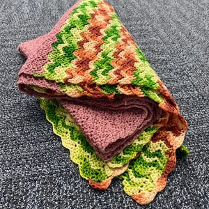 May include: A folded, handmade crochet blanket. The blanket features a scalloped edge and a chevron pattern in shades of green, orange, and yellow. The interior is a solid mauve color. The blanket is folded neatly.