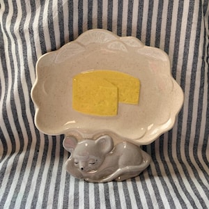 May include: A whimsical ceramic cheese and mouse dish. The off-white, scalloped-edge plate features two yellow cheese wedges. A sleeping gray mouse forms the base of the dish. The background is a striped fabric.