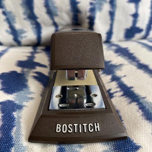 May include: A brown Bostitch stapler with a textured finish. The stapler has a silver metal base and the brand name "BOSTITCH" printed in white. The background is a blue and white patterned fabric.