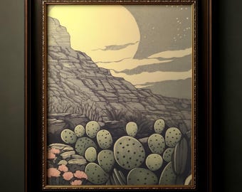May include: Framed artwork depicting a desert landscape under a large, pale yellow moon. The scene includes a mountain, cacti, and pink flowers. The artwork is in a dark brown frame.