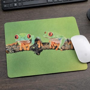 May include: A green mousepad featuring two red-eyed tree frogs perched on a branch against a green background. The frogs have bright orange feet and are facing the viewer. A white computer mouse is to the right.