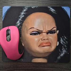 May include: A pink wireless computer mouse next to a rectangular mousepad. The mousepad features a close-up of a doll's face with a displeased expression, dark hair, and dramatic makeup. The background is black and blue.