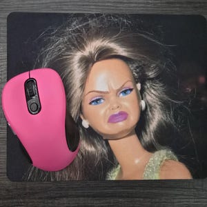 Può includere: Un tappetino per mouse rettangolare con l'immagine stampata di una bambola con lunghi capelli castani, occhi azzurri e un'espressione di disgusto con labbra viola, abbinato a un mouse per computer rosa acceso con rotellina e impugnatura nere.