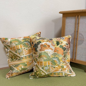 以下が含まれることがあります： 金、オレンジ、緑の色合いの、花柄模様の正方形の装飾用クッション2つ。 クッションは伝統的な日本のデザインです。 クッションは緑色のマットの上に置かれています。