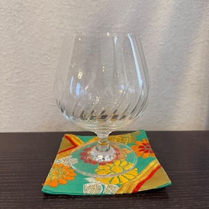 以下が含まれることがあります： スパイラルデザインの透明なブランデーグラスが、正方形のコースターの上に置かれています。コースターは、緑色の背景にオレンジと黄色の花柄と金のアクセントが施されています。グラスは空です。