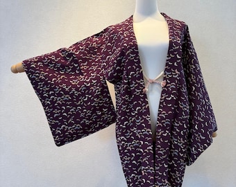 May include: A dark purple kimono-style jacket with a repeating pattern of small, crescent-shaped designs in white and gold. The jacket has wide sleeves and a decorative closure at the chest. The garment is displayed on a mannequin.