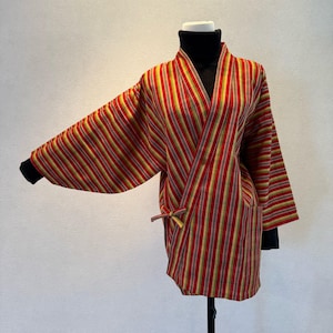 Puede incluir: Chaqueta tipo kimono a rayas rojas, amarillas y grises, con mangas anchas y diseño envolvente. La chaqueta tiene cierre de lazo y se lleva sobre un cuello alto negro. Las mangas tienen puños negros.