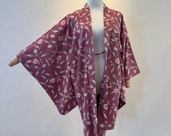 May include: A mauve-colored kimono-style robe with a floral pattern of white flowers and green leaves. The robe is displayed on a mannequin, showcasing its open front and wide sleeves. The fabric appears lightweight and suitable for layering.