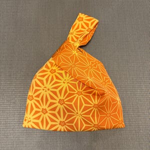 Op de afbeelding: Oranje en gele stoffen tas met een geknoopt handvat. De tas heeft een herhalend geometrisch patroon van stervormige figuren en bloemaccenten. De stof is zacht en opvouwbaar. Geschikt voor het dragen van spullen.