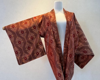 May include: A brown kimono with a repeating geometric pattern in red and yellow. The kimono is displayed on a white mannequin, showcasing its open front and wide sleeves. The fabric has a subtle sheen.