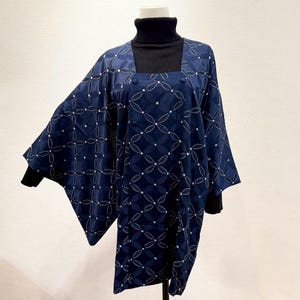 以下が含まれることがあります： ネイビーブルーの着物風トップス。白い幾何学的な花柄が特徴です。トップスはスクエアネックで、ボタンが3つ付いています。袖は幅広で流れるようなデザインです。黒のタートルネックの上に着用されています。
