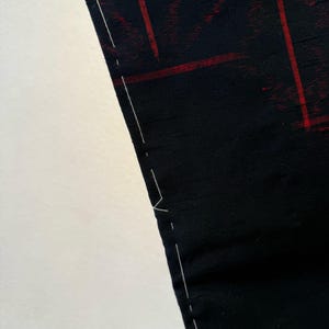 May include: Close-up of a black fabric with a red grid pattern. The fabric has a subtle sheen and is held up, revealing a white background. White stitching is visible along the edge of the fabric, suggesting a seam or hem. The overall aesthetic is modern and minimalist.