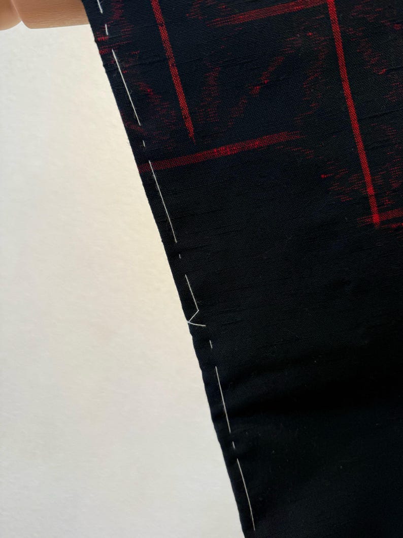 May include: Close-up of a black fabric with a red grid pattern. The fabric has a subtle sheen and is held up, revealing a white background. White stitching is visible along the edge of the fabric, suggesting a seam or hem. The overall aesthetic is modern and minimalist.