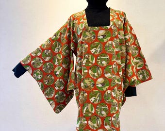 May include: A vibrant, patterned kimono-style top. The garment features a bold orange background with large, circular green and tan designs. The sleeves have a navy blue trim. The top is worn over a black turtleneck.