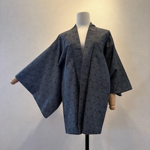Pode incluir: Um quimono azul escuro com um padrão floral repetido. O quimono é exibido em um manequim com braços de madeira. A peça tem mangas largas e um design de decote em V. O tecido parece ser um material macio e tecido.