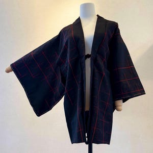 May include: A black kimono-style jacket with a red plaid pattern. The jacket has wide sleeves and a draped front, displayed on a mannequin. The fabric appears to be a lightweight material, suitable for layering or warmer weather.