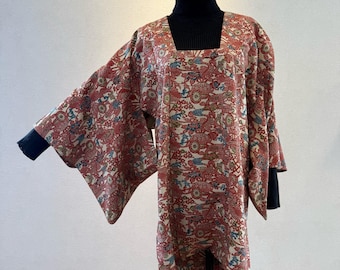 May include: A patterned kimono-style top with a square neckline. The fabric features a red, cream, and blue floral design. The sleeves are wide and the garment is worn over a black turtleneck. The kimono is displayed on a mannequin.