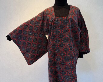May include: A red and black patterned kimono-style garment with wide sleeves. The fabric features a geometric design. The garment is worn over a black turtleneck. The kimono is displayed on a stand.