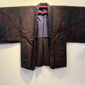 May include: Black kimono with a red grid pattern. The kimono is hanging on a red hanger against a white wall. The interior lining is a solid blue-purple color.