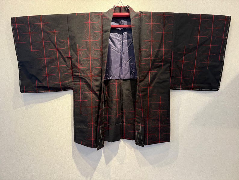 May include: Black kimono with a red grid pattern. The kimono is hanging on a red hanger against a white wall. The interior lining is a solid blue-purple color.