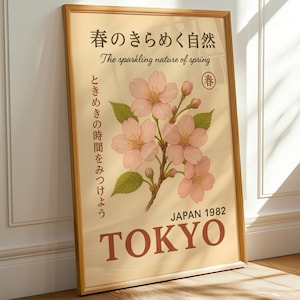 Op de afbeelding: Een ingelijste poster met een vintage esthetiek. De poster toont een bloemenillustratie van roze kersenbloesems met groene bladeren. De tekst in het Engels en Japans luidt "The sparkling nature of spring" en "TOKYO JAPAN 1982".