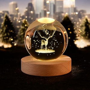 May include: A glass sphere with a 3D etched design of a deer standing in a field with stars. The sphere is illuminated from within and sits on a wooden base.