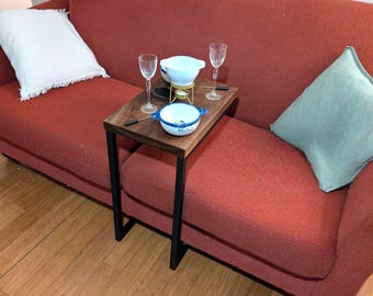 Puede incluir: Una bandeja de servir de madera con patas de metal negro se encuentra sobre un sofá rojo. La bandeja contiene una olla de fondue blanca con un soporte dorado, dos copas de vino y un pequeño tazón azul y blanco.