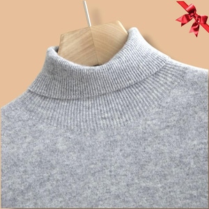 May include: A close-up of a light gray turtleneck sweater. The sweater is made of a soft, ribbed knit material. The turtleneck is folded over and the sweater is hanging on a wooden hanger.