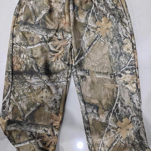 May include: Camouflage-patterned pants in shades of brown, green, and tan. The pants feature a realistic forest design with tree branches and leaves. The pants have an elastic waistband and a relaxed fit, suitable for outdoor activities.
