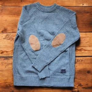 May include: A blue knitted jumper with brown leather elbow patches. The jumper has a ribbed texture and a crew neck.