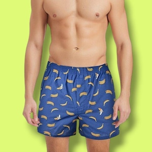 May include: Blue boxer shorts with a repeating pattern of yellow bananas. The shorts have an elastic waistband and are made of a lightweight fabric. The background is a solid lime green.