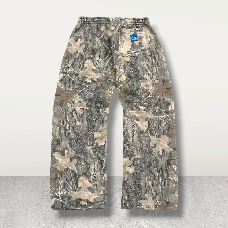 May include: Camouflage-patterned sweatpants with an elastic waistband and a small blue tag on the back. The pants feature a realistic tree bark and leaf design in shades of brown, green, and tan. The relaxed fit and neutral colors make them suitable for outdoor activities.