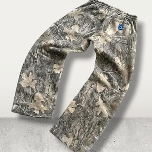 May include: Camouflage-patterned sweatpants in shades of green, brown, and beige. The pants feature an elastic waistband and a back pocket with a blue logo. The fabric has a print of leaves and tree bark.
