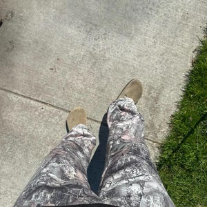 May include: A person wearing camouflage pants with a black shirt and tan suede shoes. The pants have a gray, white, and brown pattern. The person is standing on a concrete surface next to green grass.
